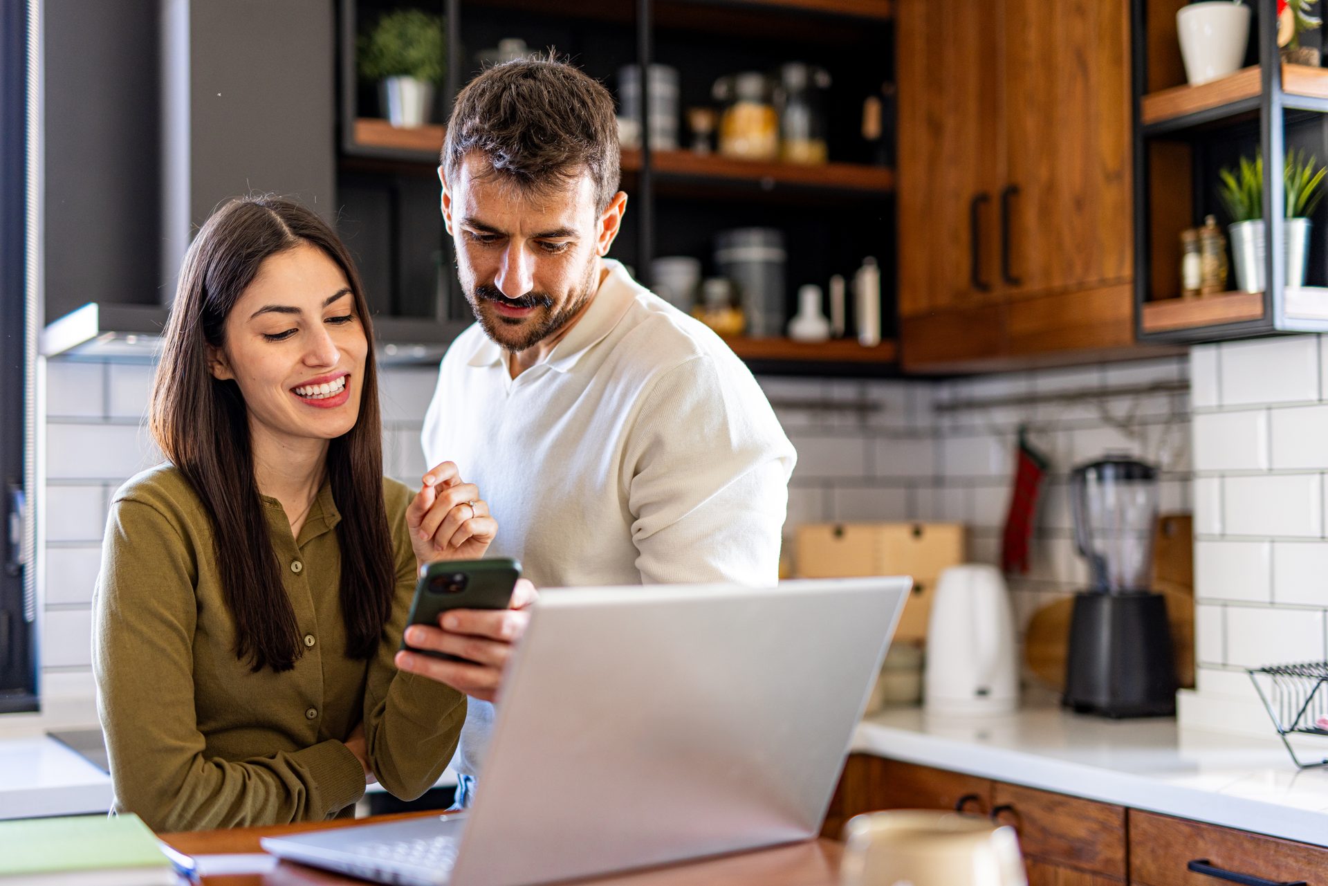 Couple smiling over a phone and laptop while planning their savings at the kitchen table