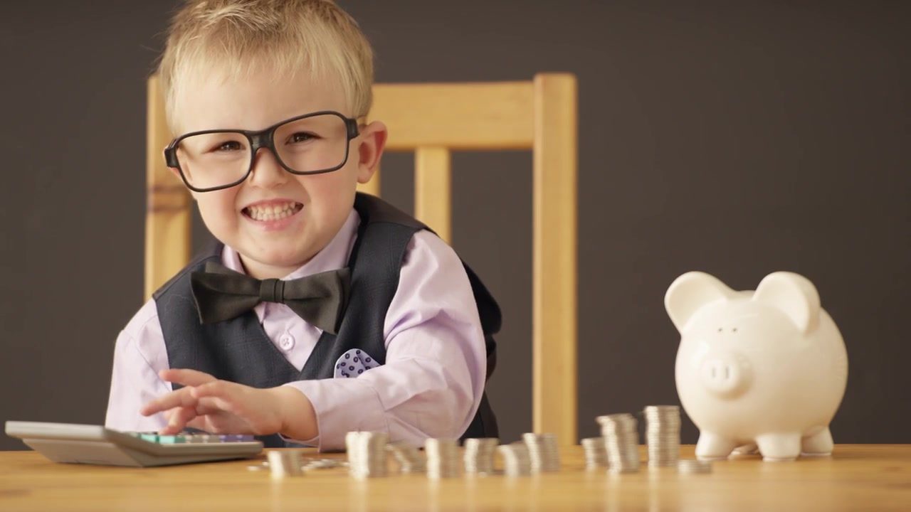 Smiling young boy in glasses and a bow tie with a calculator, stacks of coins, and a piggy bank
