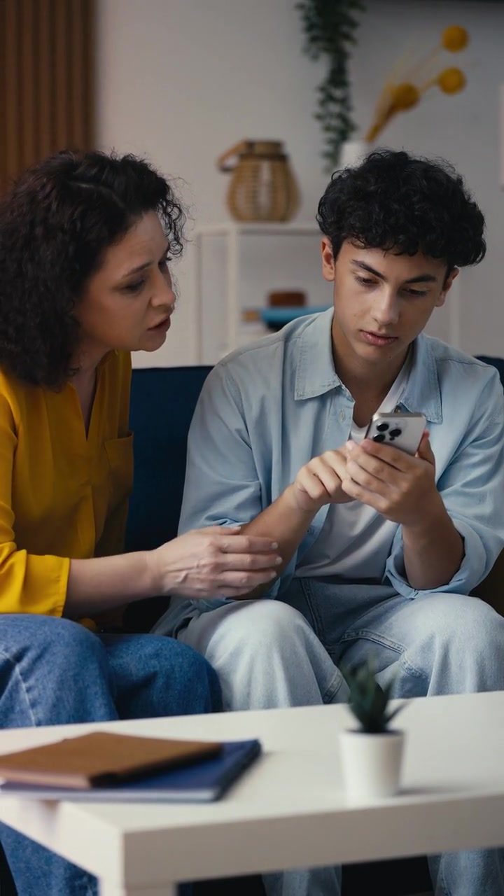 Teen showing his mother a banking app on his phone while they sit together on a couch