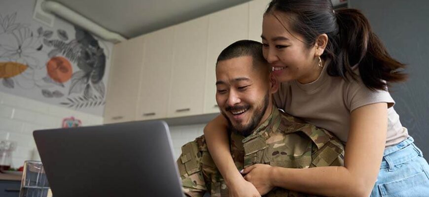 a woman wraps her arms around her military husband as he begins the process of opening a checking account online